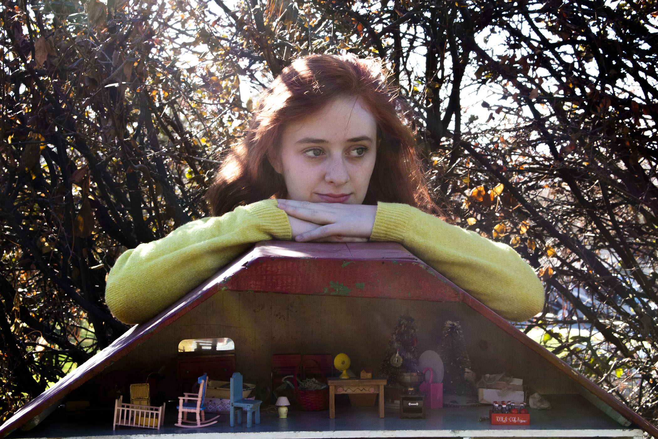 A red-haired woman sits outside, looking outwards as her hands and head rest upon a red-roofed dollhouse filled with colorful, miniature furniture of all sorts