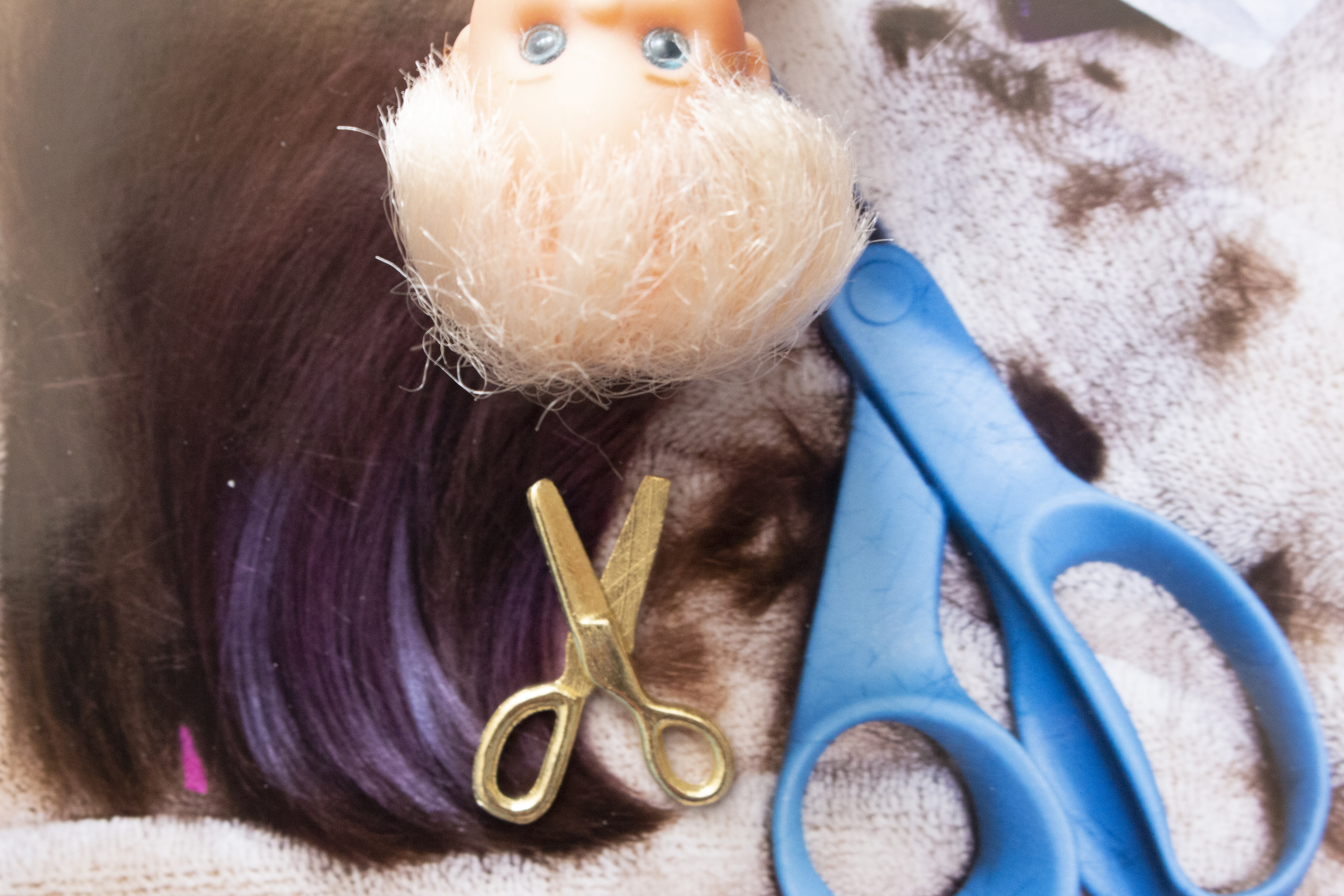 An upside down doll head and a miniature pair of scissors float over a background of a pair of scissors and cut hair