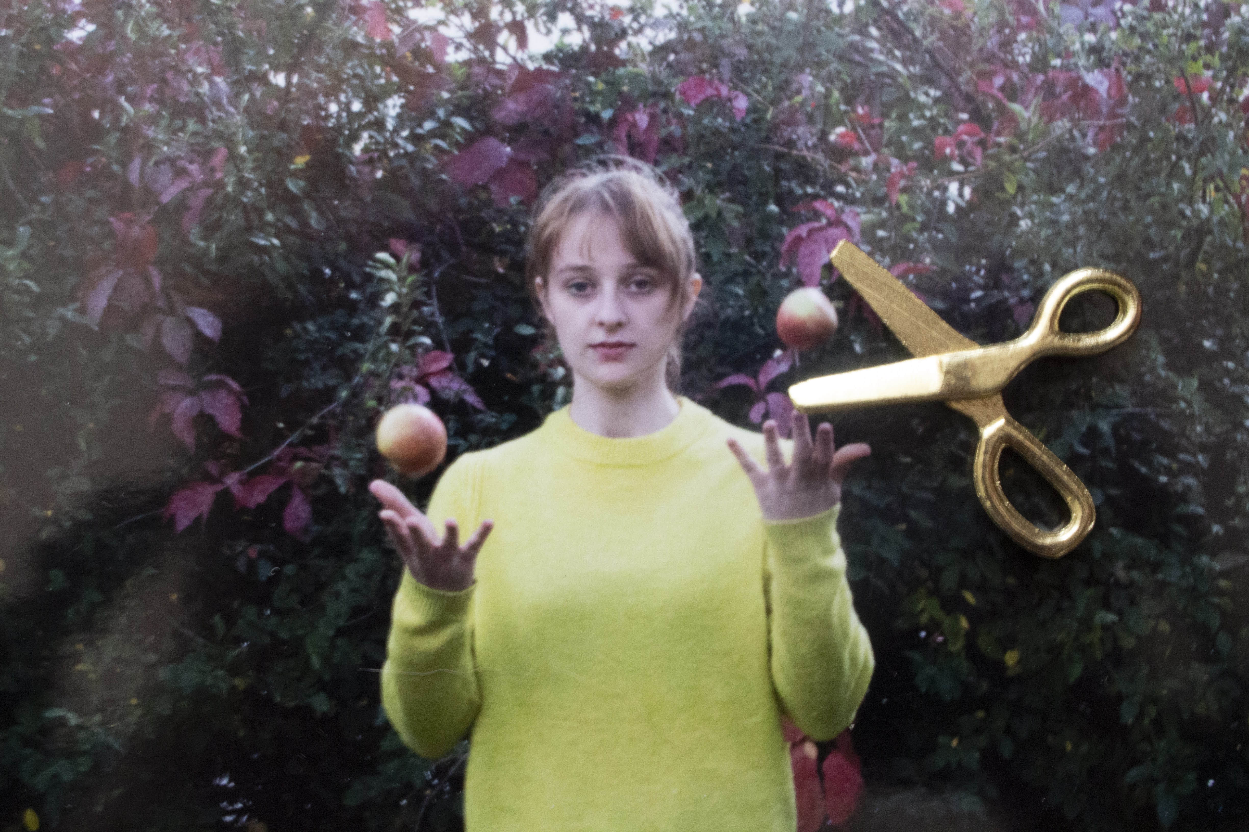 A blonde woman in a yellow sweater is outside, throwing apples up into the air. They are frozen next to her in the photograph. On top of the photograph, a miniature pair of golden scissors sits, looking as if it is about to cut one of the apples.