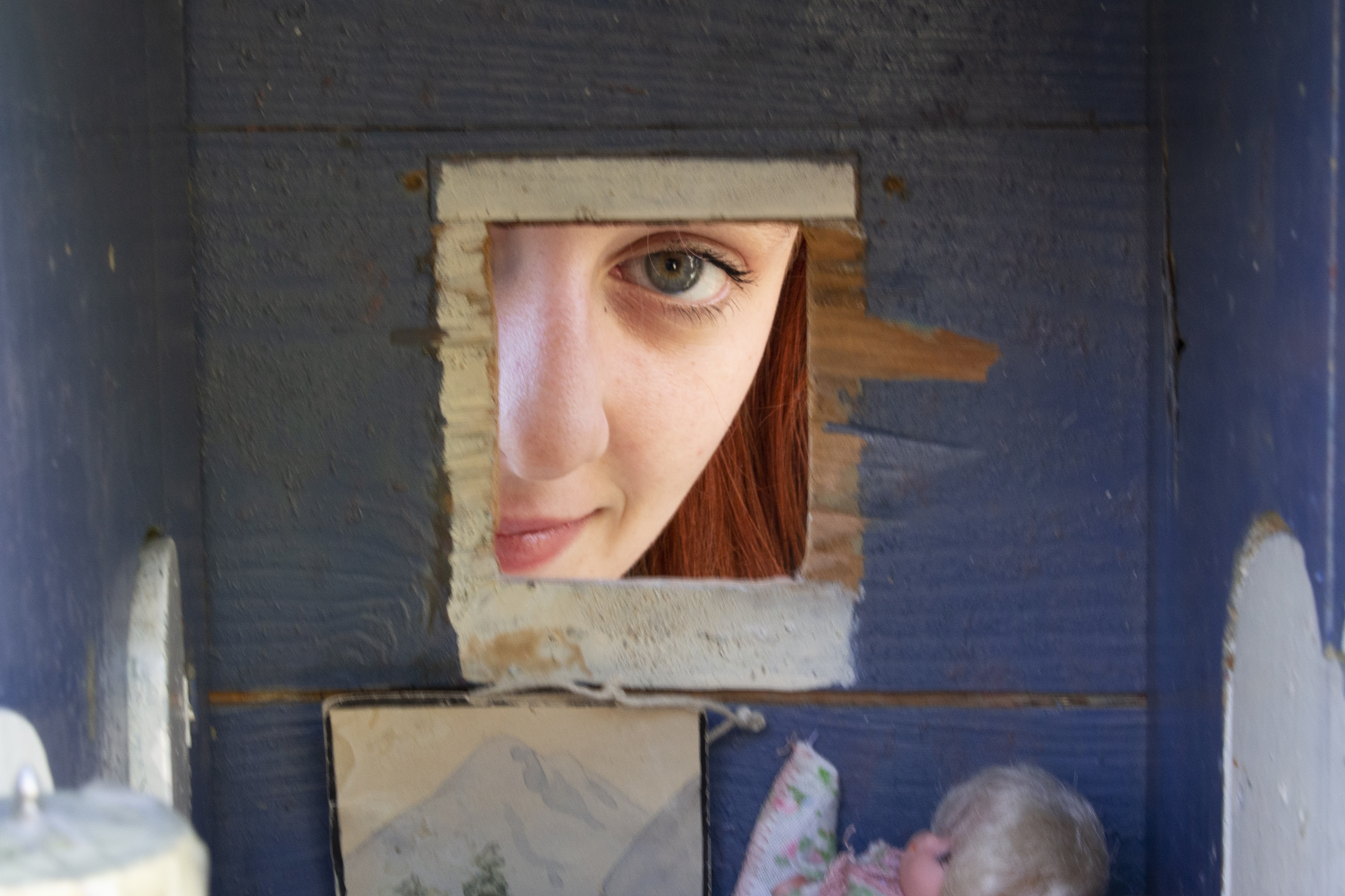 A red-haired woman peers through the window of a blue-walled dollhouse from the outside. Only her eye and a portion of her nose are visible. Below her, in the dollhouse, a doll sleeps in a bed.